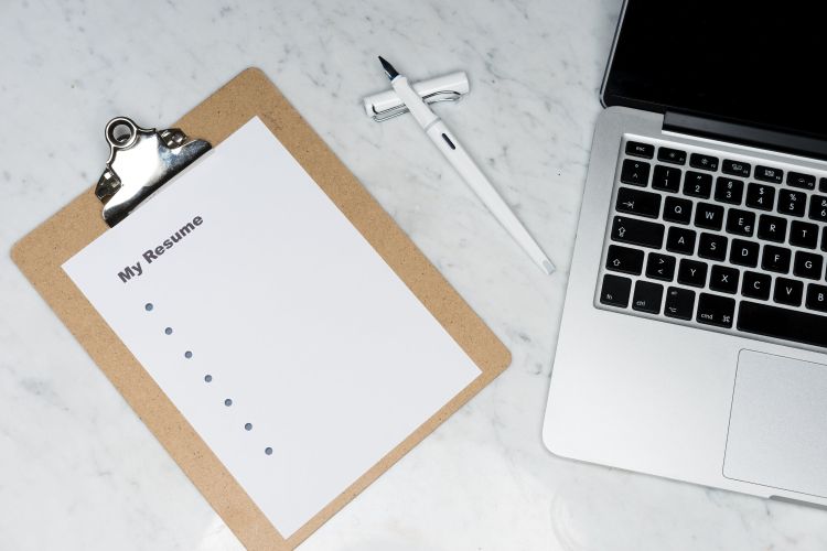 A clipboard next to a computer and pen. On the clipboard is a piece of paper that says my resume.