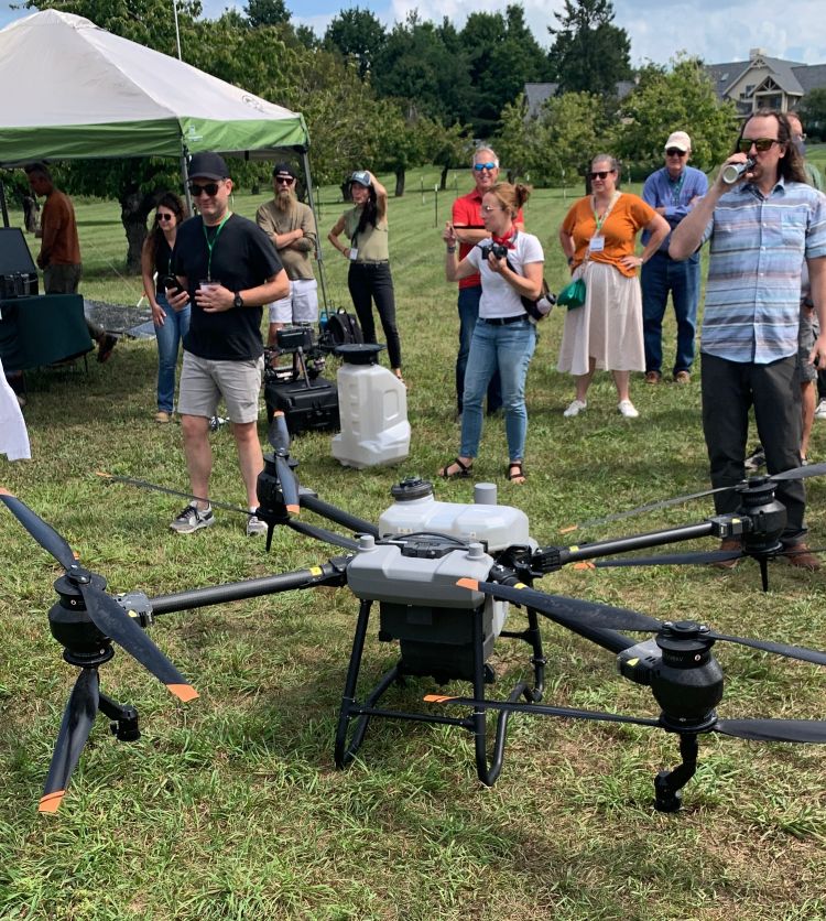 A large agricultural drone with multiple rotors sits on grass during an outdoor demonstration, while a group of adults stands nearby watching.