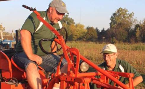 Farmer on a tractor with someone kneeling in the field beside the tractor.