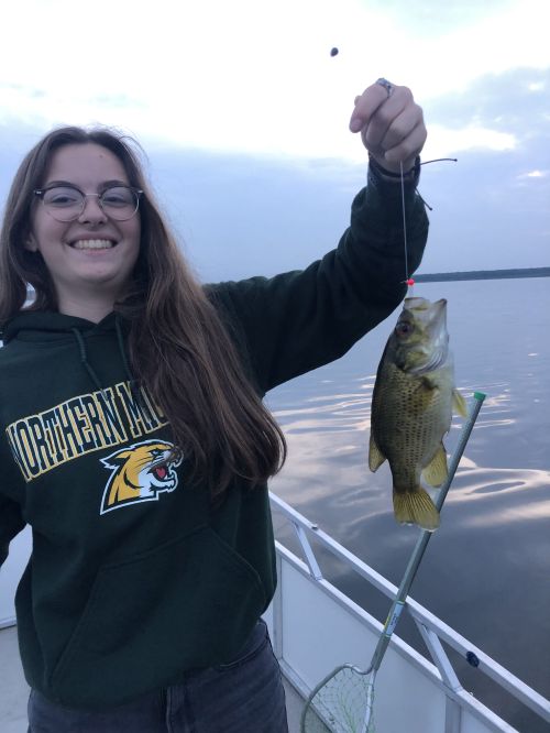 A girl with dark hair and glasses in a dark NMU sweatshirt holding up a fish.