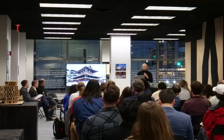 An image of the back of an audience sitting in the Tall Timber exhibit space, all facing a panel of speakers and a tv monitor displaying an image of a building.