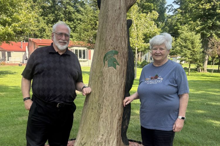 J. Mark  and Kathy Scriber posing in front of a tree with an MSU Spartan helmet logo carved into it.