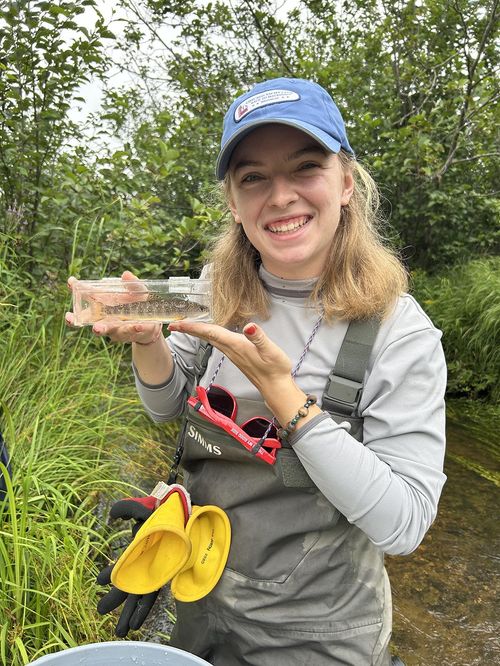 Woman wearing a overalls and a hat, stands in a stream and holds a clear sampling box and a small fish.
