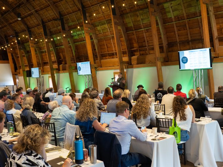 Attendees of the Dirt to Glass conference sit at tables inside a large cathedral barn, watching someone give a presentation at a podium at the front of the room. Lights dangle from the curved ceiling, and three TVs on the wall project a PowerPoint presentation.