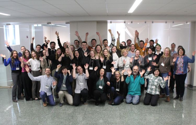 A large group of symposium participants stands together indoors in a bright, modern building, facing the camera with raised hands. The group represents students, faculty, and attendees of the 2025 FW GSO Symposium.