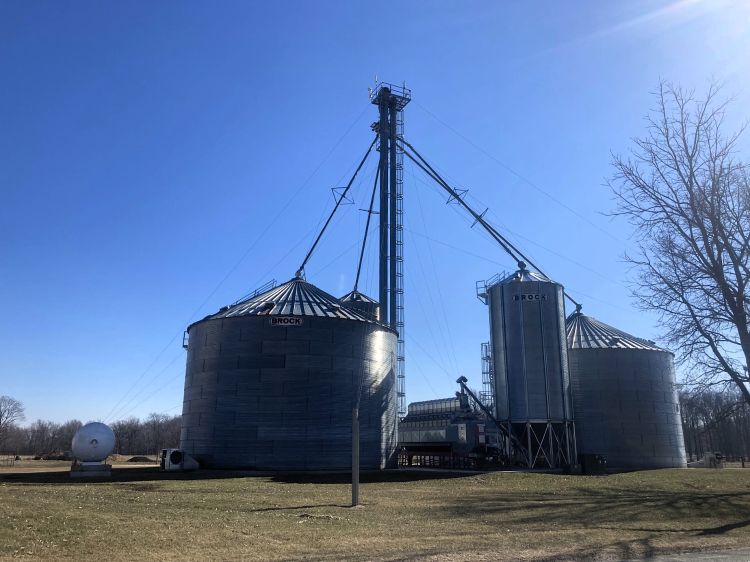 A farm grain storage system with several large silver metal silos labeled “Brock,” connected by tall conveyors and a central vertical structure used for transferring grain. The silos stand on a grassy area under a clear blue sky, with a leafless tree on the right and a white propane tank on the left.