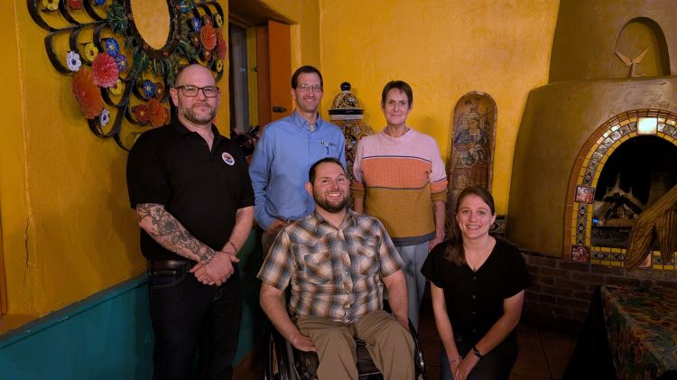Group photo of five adults indoors, smiling and posed against a warm yellow wall with decorative art and a fireplace. One person is seated in a wheelchair in the center, with others standing and kneeling nearby.