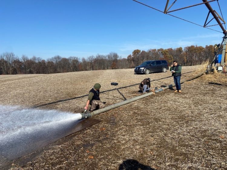 Three people stand in a harvested field working on a large irrigation pipe connected to a pivot system, with water forcefully spraying from the open end while a vehicle is parked nearby under a clear blue sky.