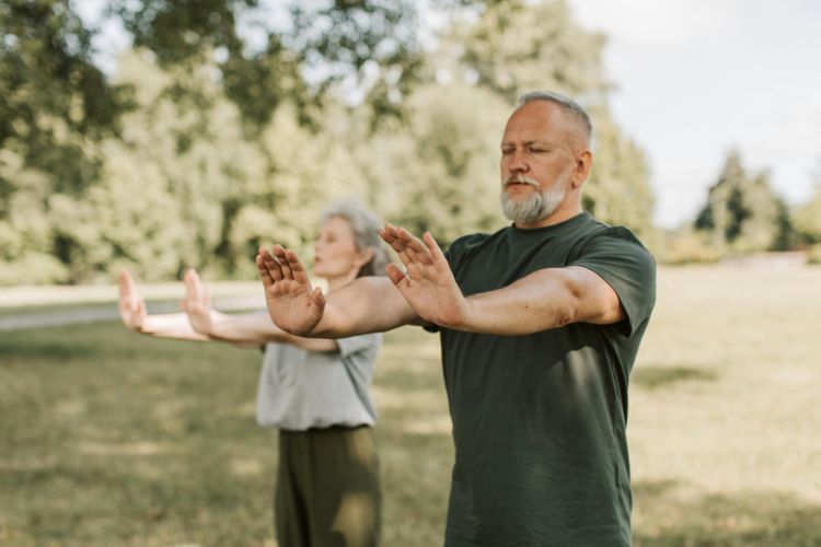 Two older adults practice meditation outside, they hold their hands outstretched.
