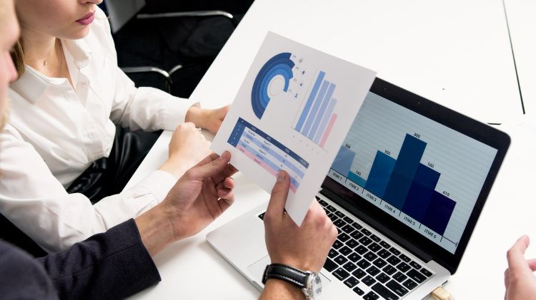 Two individuals reviewing charts and graphs on paper handout and laptop screen.