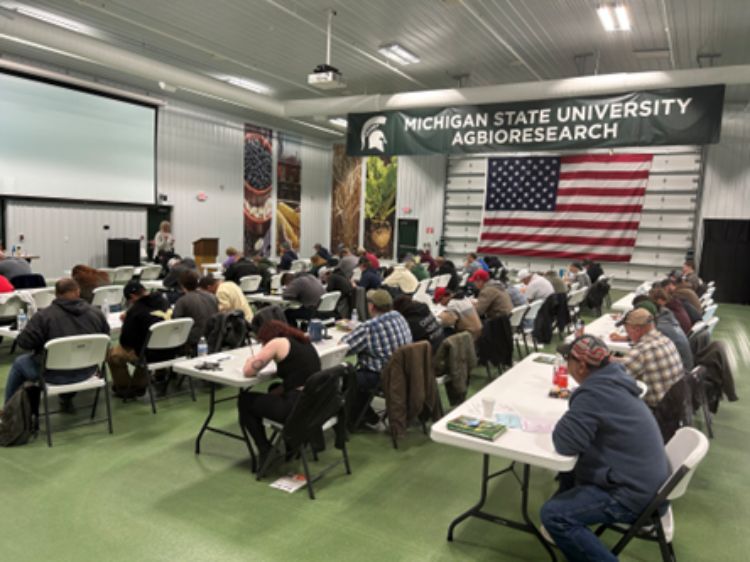 A large group of people seated at tables in a bright meeting hall at Michigan State University AgBioResearch facility. Attendees face a presenter near the front, with notebooks, papers and drinks on the tables.