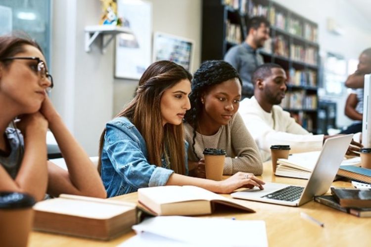 Two women looking intently at a computer screen in a library setting surrounded by others studying.