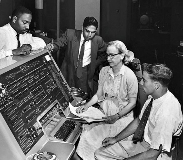 A historical black and white photo of a woman and three men staring at a large computer.