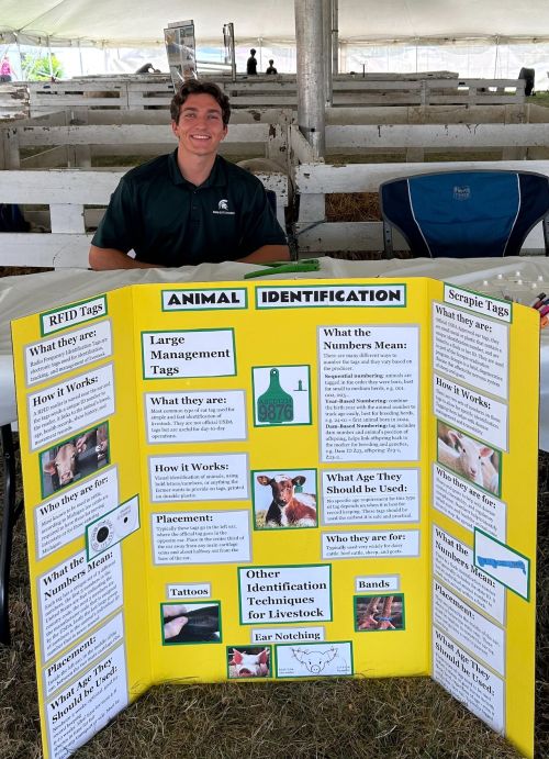 A young man in a green shirt behind a yellow poster board with information about animal identification.