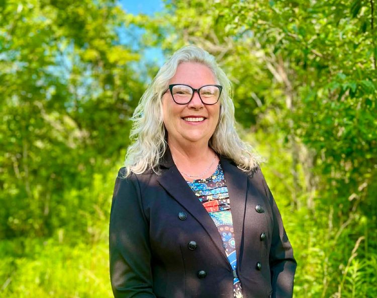 A woman in a dark blazer with light hair and classes in an outdoor setting.