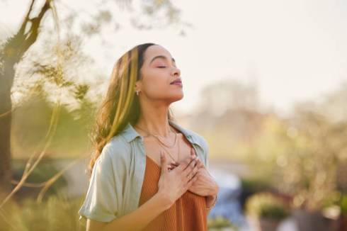 A female sitting down outside on the grass with her eyes closed and her hands over her chest.