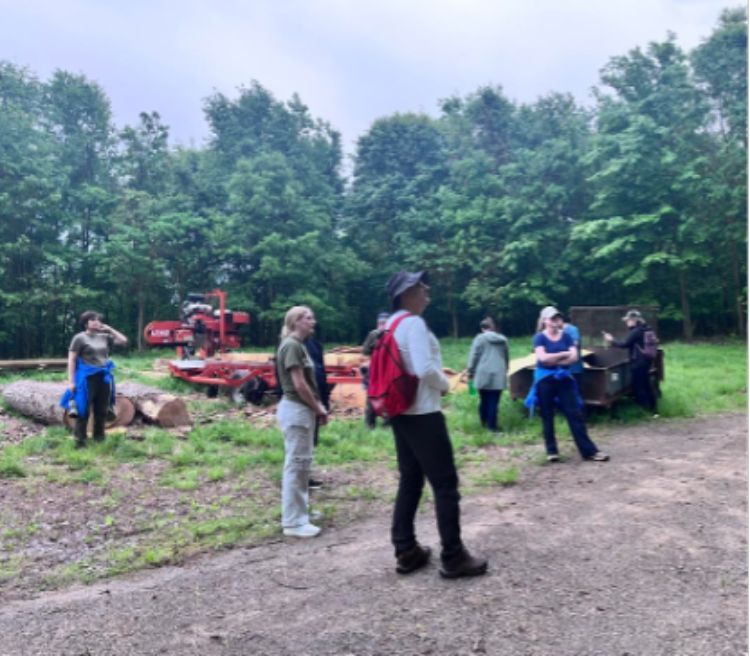 Photograph of a group of seven people standing outdoors near forestry equipment and cut logs, surrounded by dense green trees.