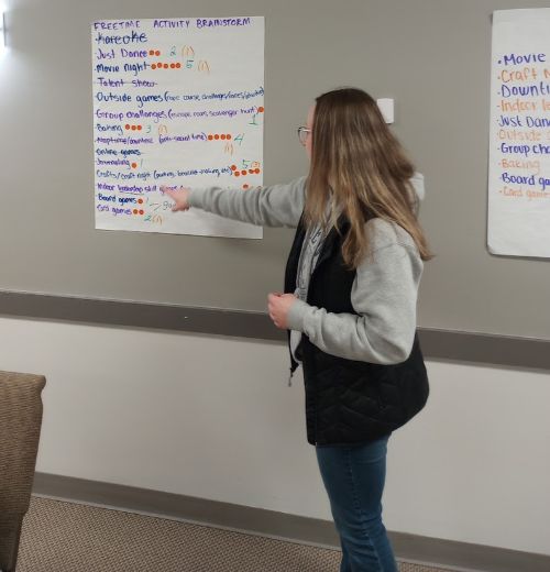 A young person pointing to a list of ideas written on a large sticky note and posted on the wall.