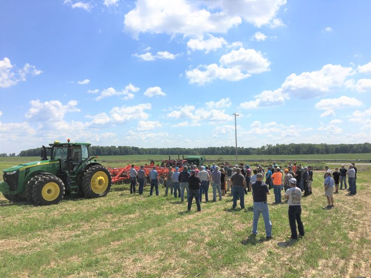 people stand and look at a red roller crimper behind a green tractor on green grass
