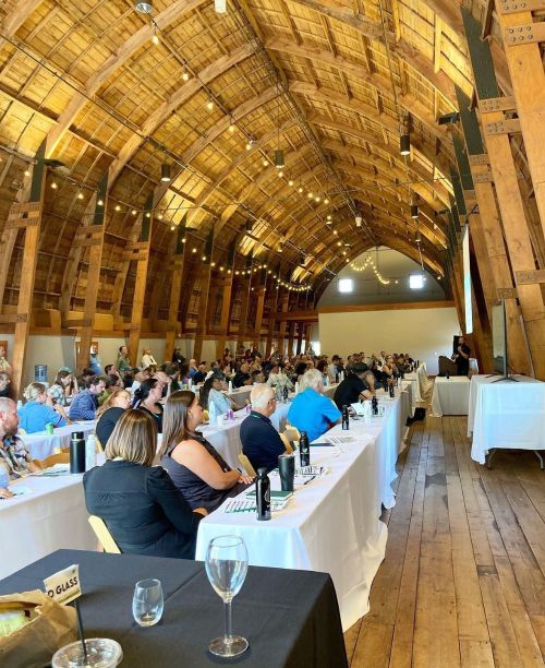 Attendees sit at long tables inside a timber-framed barn, listening to a speaker at the front during a vineyard and wine conference, with string lights overhead and presentation materials on the tables.