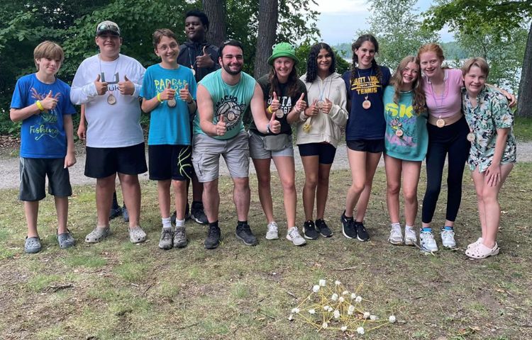 a group of youth standing in a line with thumbs up and arms around each other. At their feet is a tower made of our spaghetti and marshmallows.