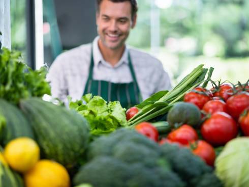Man with a green apron standing behind a variety of vegetables