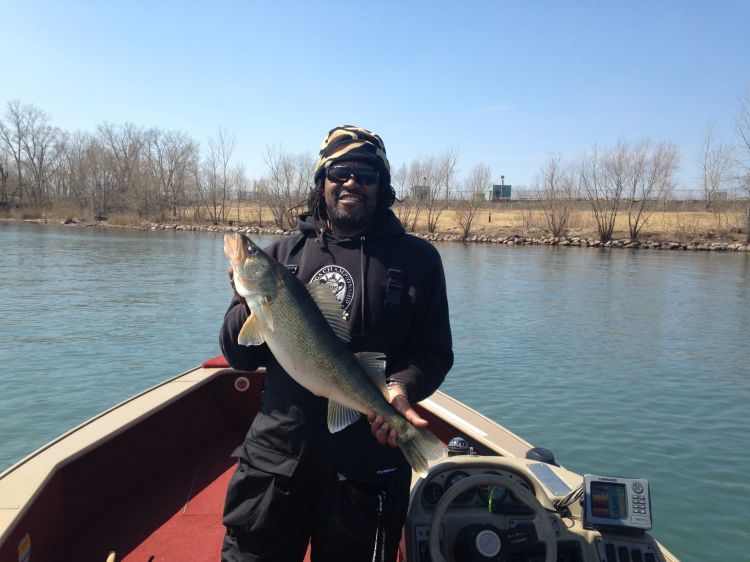 A man in a boat on the water holds a walleye fish while smiling and facing the camera. The shoreline is seen in the background.