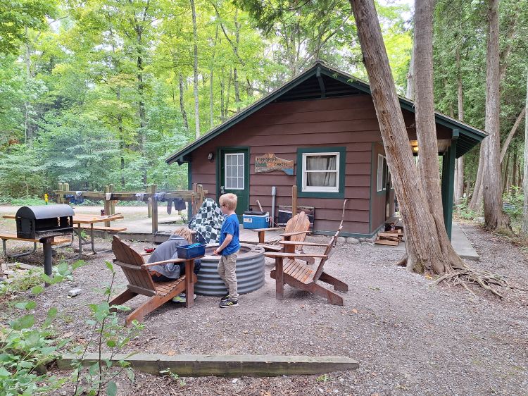 Two young boys sitting next to a campfire beside a brown cabin in the woods.