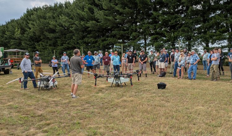 A man speaks to a group of about 30 adults gathered in a grassy field while standing beside two large agricultural drones equipped with spray tanks and multiple rotors.