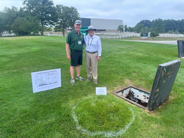 Two gentleman stand next to a hole in the ground with an open lid with a sign with a graph on it in the foreground.
