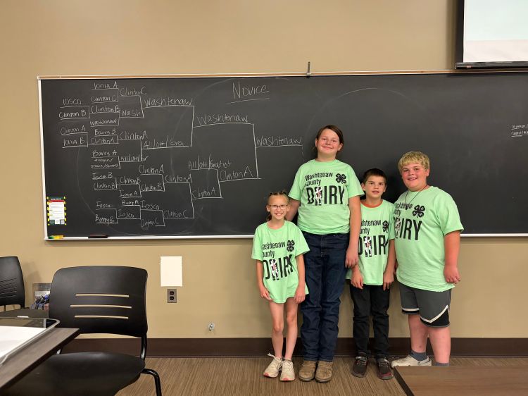 A group of youth in matching tshirts standing in front of a bracket on a black board.