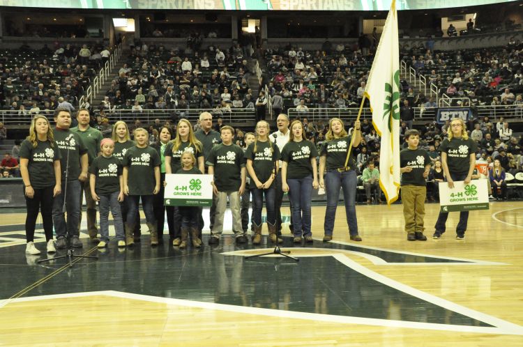 A group of youth dressed in green and white 4-H shirts at center court at the Breslin center. Holding 4-H flag and 4-H Grows Here signs.