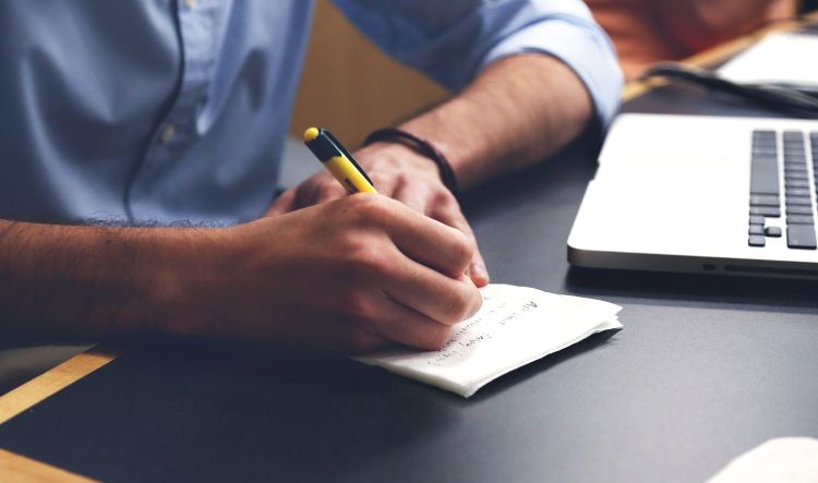 Man in blue button-down shirt writing with yellow pen on a piece of paper next to a laptop.