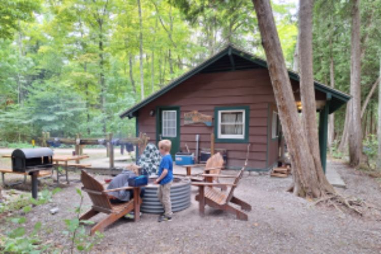 Two young boys sitting next to a campfire beside a brown cabin in the woods.