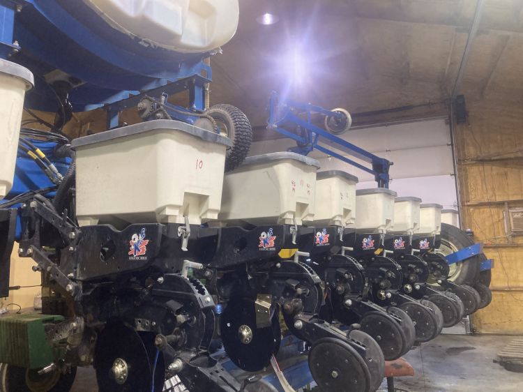 A row-crop planter with multiple white seed hoppers is parked inside a farm workshop.