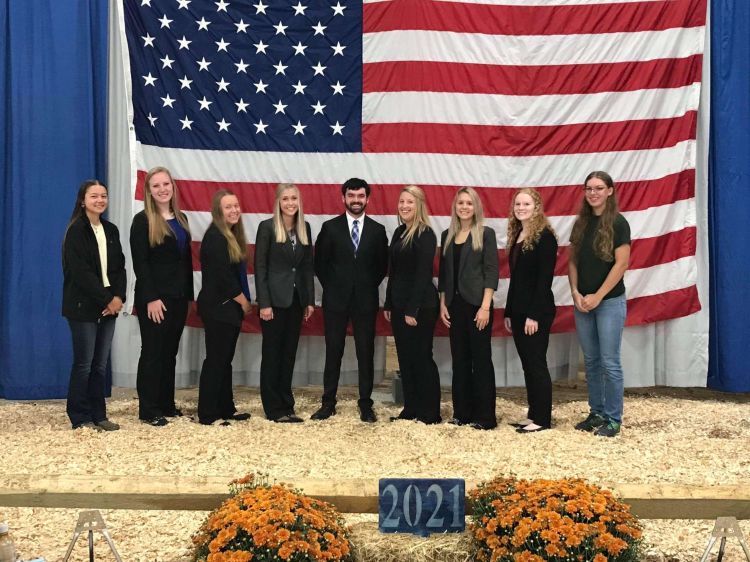 A group of 9 people posing together in front of the American flag.