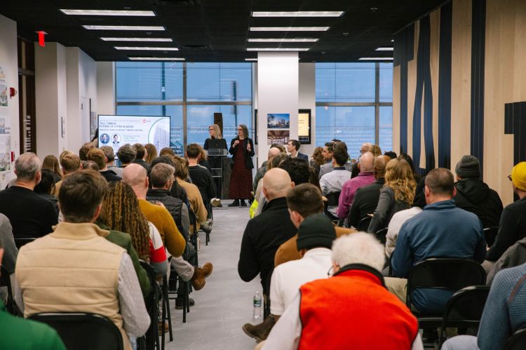 An audience of people gathered, sitting in chairs in rows facing a presenter with slides displayed on a tv screen.