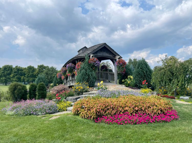 A scenic garden landscape featuring a rustic wooden gazebo surrounded by colorful flower beds. The beds are filled with pink, purple, yellow, and red flowers, with manicured shrubs and trees in the background under a partly cloudy sky.