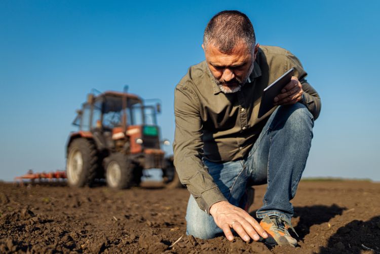 A farmer, holding a tablet in one hand, bends down on a knee in an open field touching the soil beneath him. Behind him is a parked tractor.