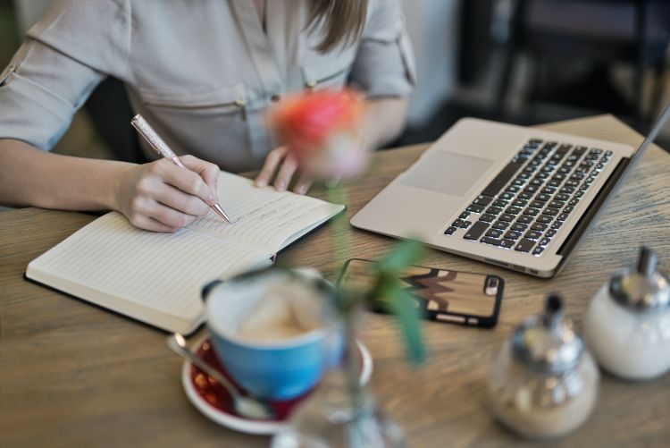 A person takes notes in a notepad while looking at their computer.