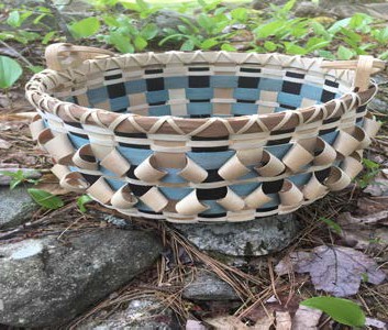 Handwoven decorative ash basket with black and natural wood patterns resting on rocks in a forest setting.
