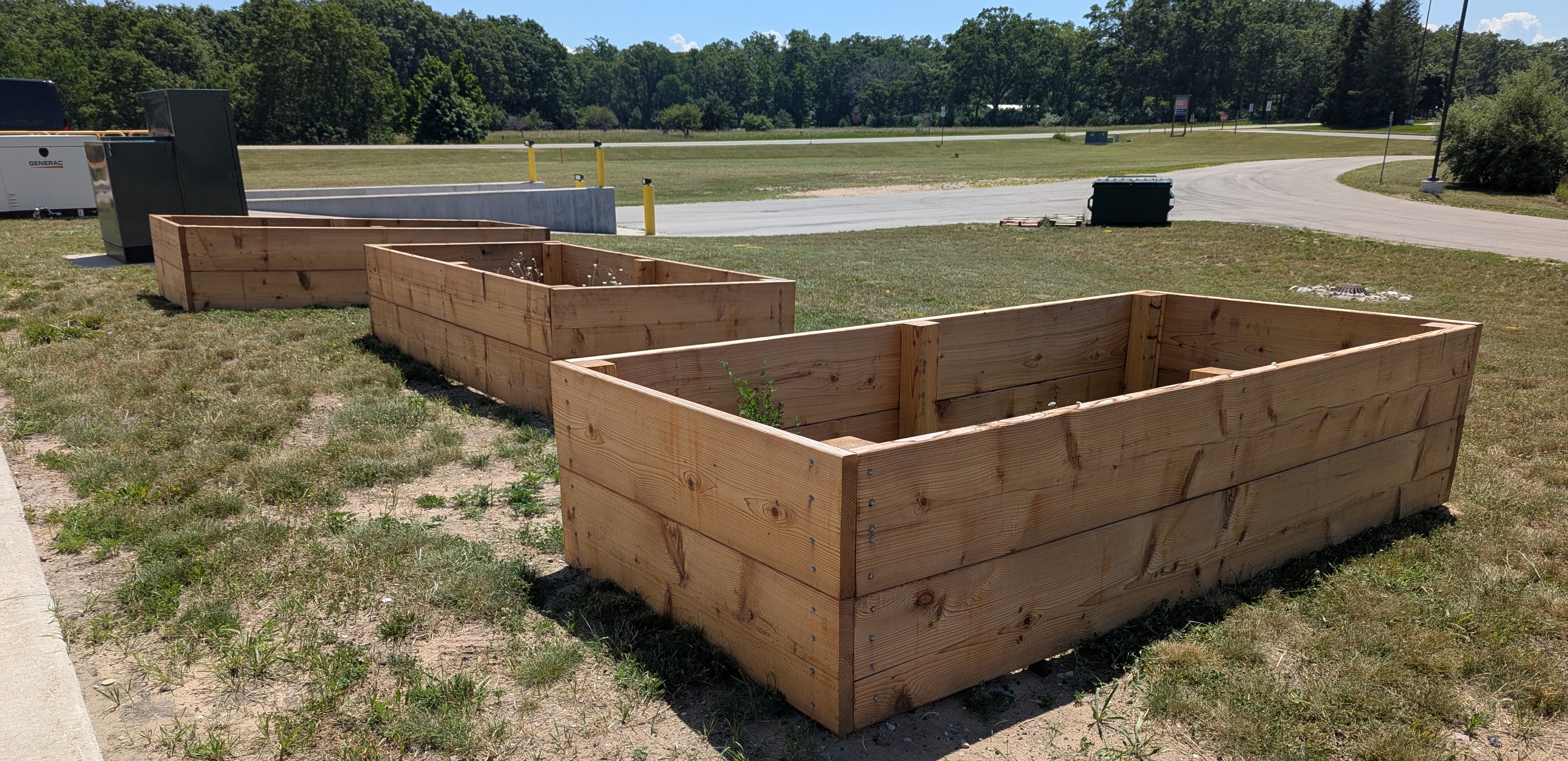 Three wooden raised garden beds on grassy ground with a road and trees in the background.