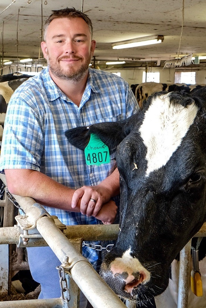 Adam Lock standing inside a dairy barn, leaning on a metal rail next to a black‑and‑white dairy cow with a green ear tag. Other cows and barn equipment are visible in the background under overhead lights