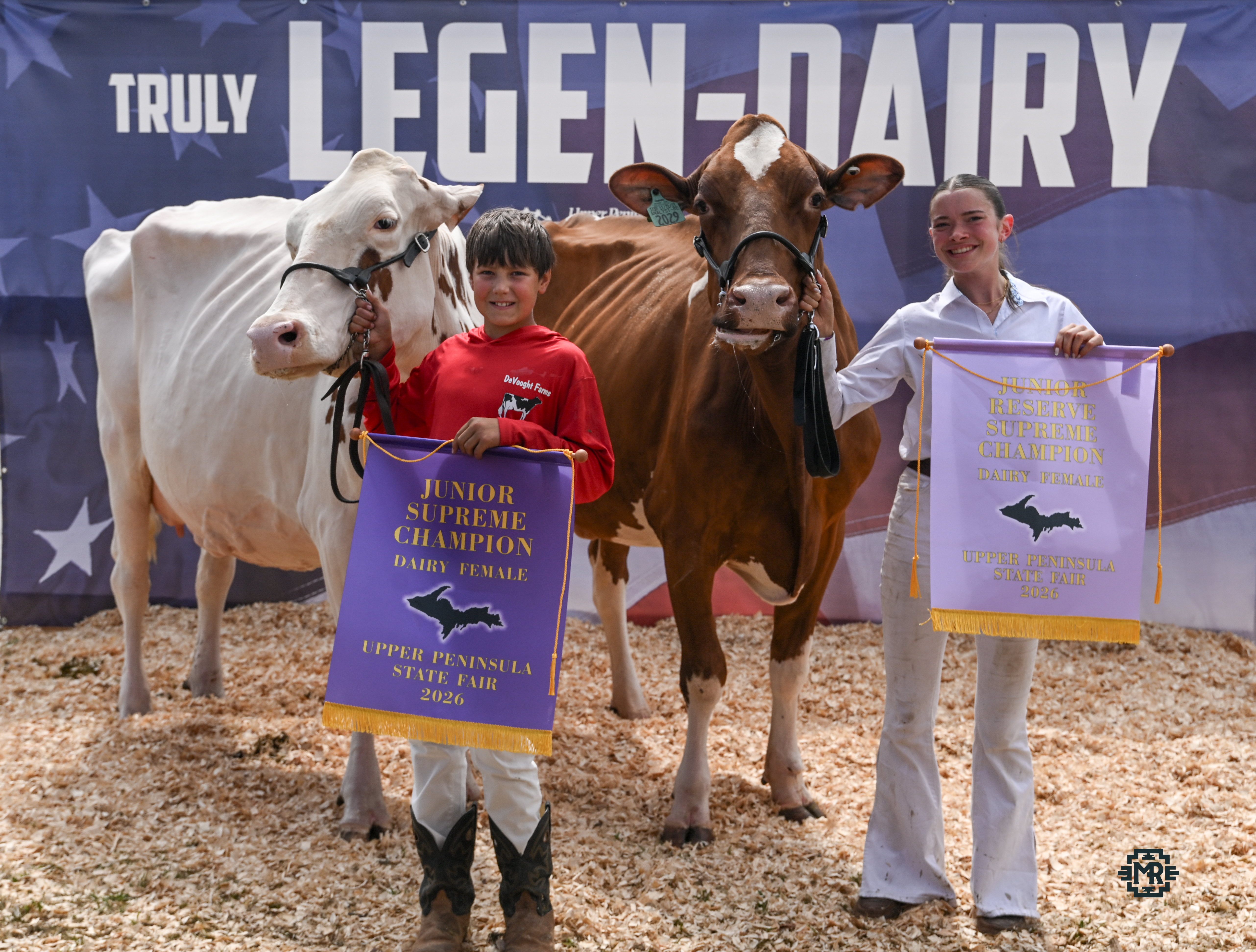 Two youth with two cows, one red and one white, holding up their championship banners in front of a backdrop.