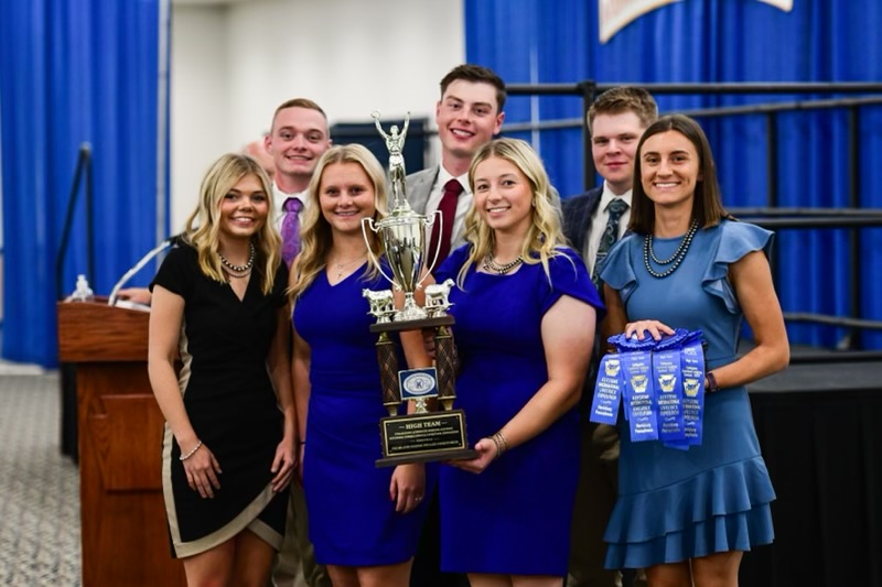 Members of the Michigan State University livestock judging team stand together indoors, holding a large trophy and multiple blue award ribbons