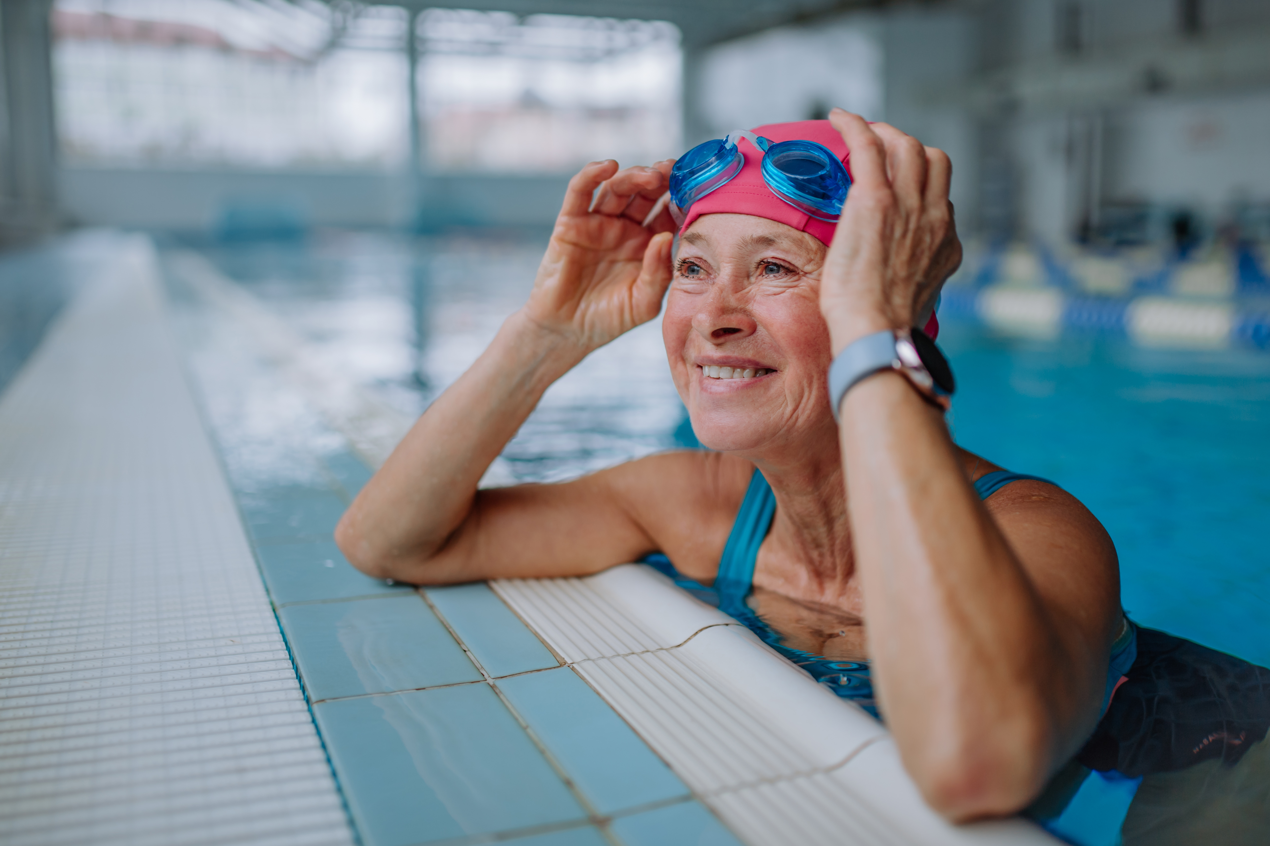 An older adult rests against the side of the pool, raising her googles up and smiling.