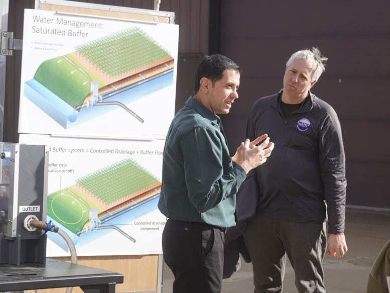 Two men stand outdoors discussing a display board about water management and saturated buffer systems. One man gestures while speaking, and the other listens attentively. The display shows diagrams of a field with drainage and buffer flow components, and equipment with an outlet pipe is visible nearby.